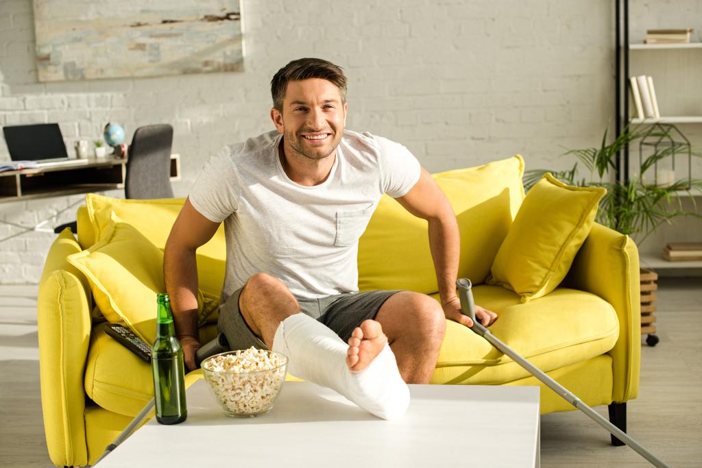 Usmívající se muž se zlomenou nohou při pohledu na kameru u pivní láhve a popcorn na konferenčním stolku v obývacím pokoji - Fotografie, Obrázek
