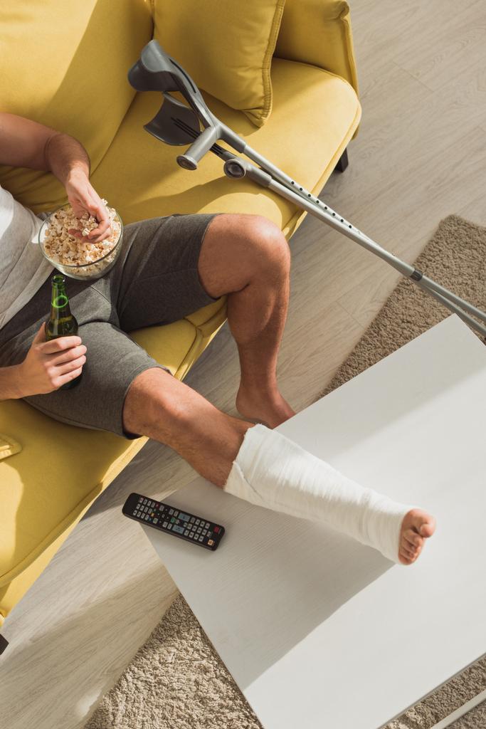 Вид сверху на человека со сломанной ногой на журнальном столике, который пьет пиво и ест кукурузу на диване дома
 - Фото, изображение