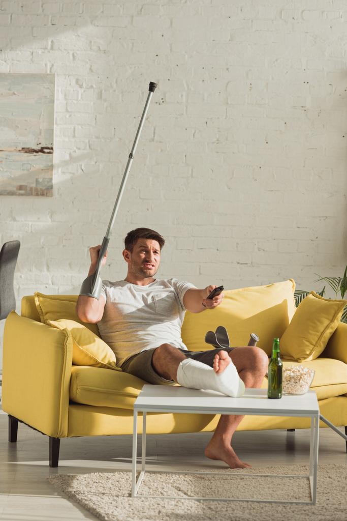 Vihainen mies, jolla on murtunut jalka katsomassa televisiota lähellä olutpulloa ja popcornia sohvapöydällä kotona
 - Valokuva, kuva