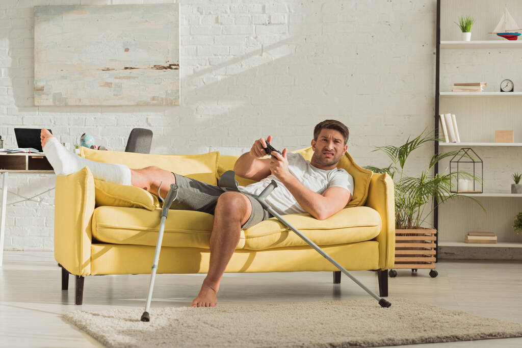 KYIV, UCRANIA - 21 de enero de 2020: Hombre molesto con la pierna rota jugando videojuegos en el sofá en la sala de estar
 - Foto, Imagen