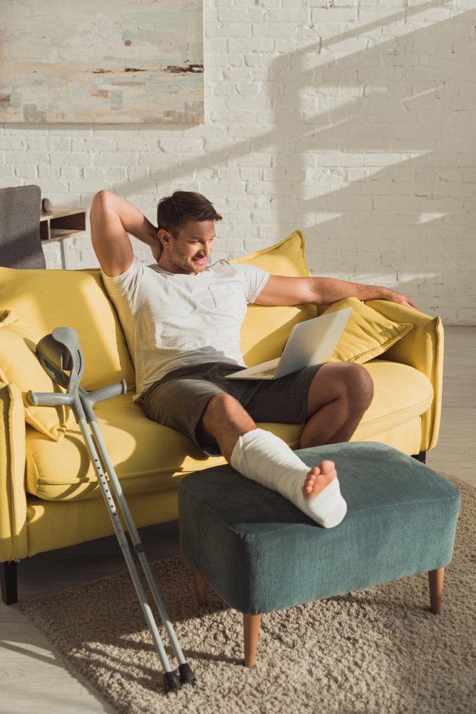 Улыбающийся мужчина со сломанной ногой на оттомане смотрит на ноутбук на диване дома
 - Фото, изображение