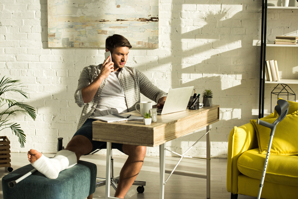 Freelancer bonito com perna quebrada usando laptop e falando no smartphone à mesa na sala de estar
 - Foto, Imagem