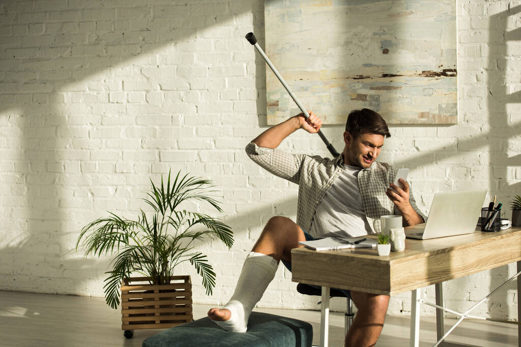 Улыбающийся мужчина со сломанной ногой держит костыль и смартфон белый сидит за столом с ноутбуком
 - Фото, изображение