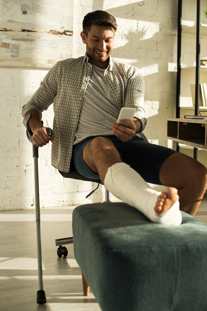 Selective focus of smiling man with broken leg using smartphone in living room - Fotoğraf, Görsel