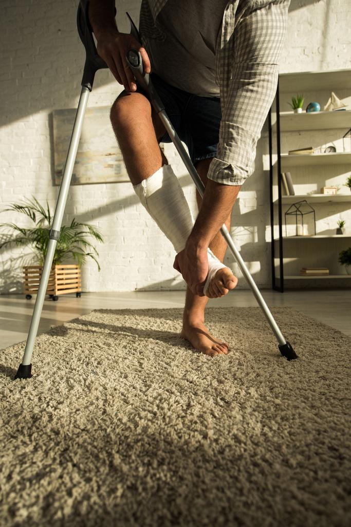 Gebonden zicht op de man die gebroken been aanraakt en krukken vasthoudt in de woonkamer - Foto, afbeelding