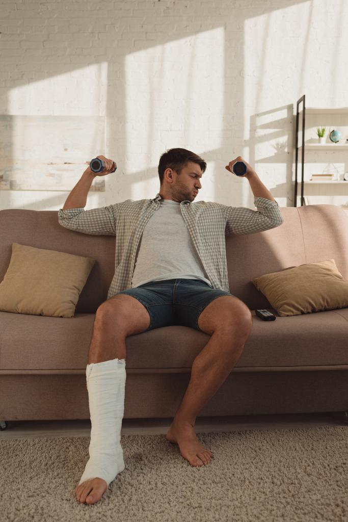 Handsome man with broken leg training with dumbbells on couch in living room - 写真・画像