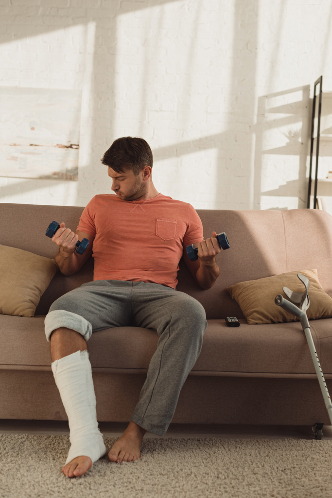 Красивый мужчина со сломанной ногой тренируется с гантелями на диване дома
 - Фото, изображение