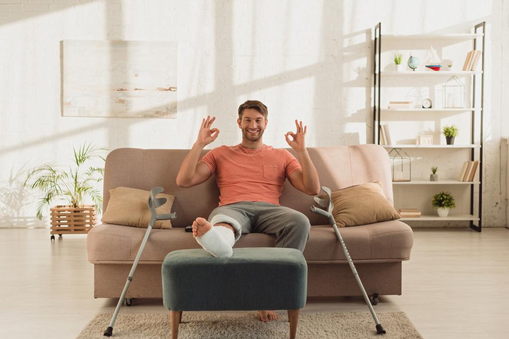Schöner Mann mit gebrochenem Bein lächelt in die Kamera und zeigt ok Zeichen auf der Couch - Foto, Bild