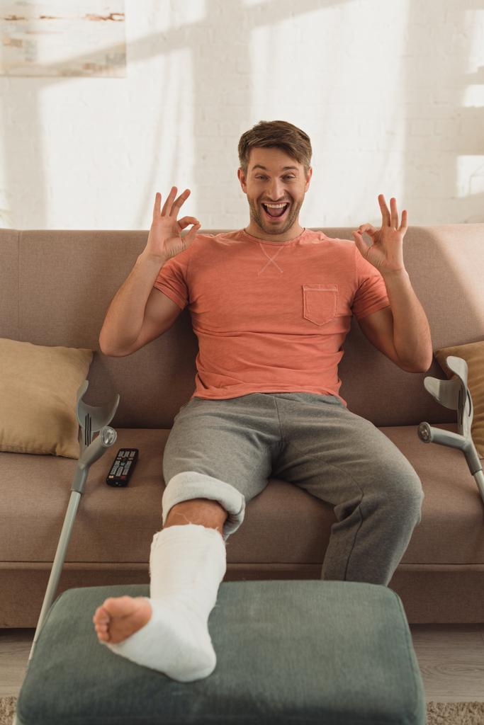 aufgebrachter Mann mit gebrochenem Bein auf Hocker zeigt Okay-Zeichen auf Sofa zu Hause - Foto, Bild
