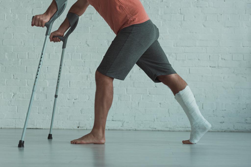 Vista lateral do homem com a perna quebrada em gesso segurando muletas em casa
 - Foto, Imagem