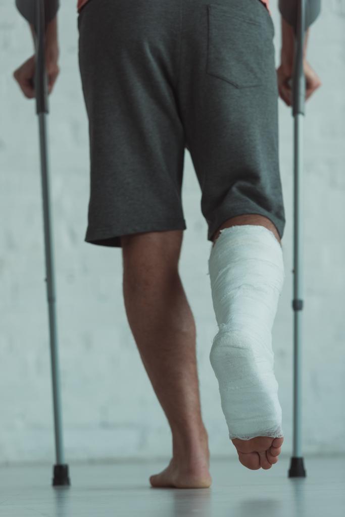 Visão traseira do homem com a perna em bandagem de gesso andando com muletas
 - Foto, Imagem