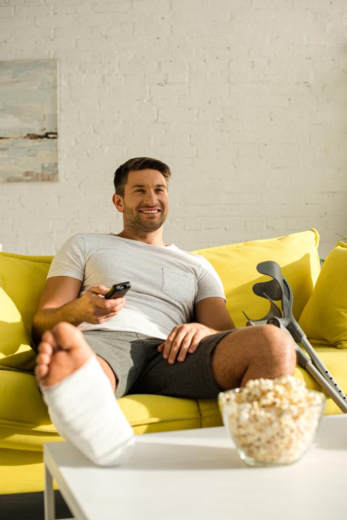 Focus selettivo dell'uomo sorridente con gamba in benda di gesso che guarda la TV vicino ai popcorn sul tavolino
 - Foto, immagini