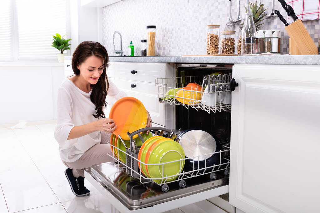 Улыбающаяся молодая женщина раскладывает тарелки в посудомоечной машине дома
 - Фото, изображение