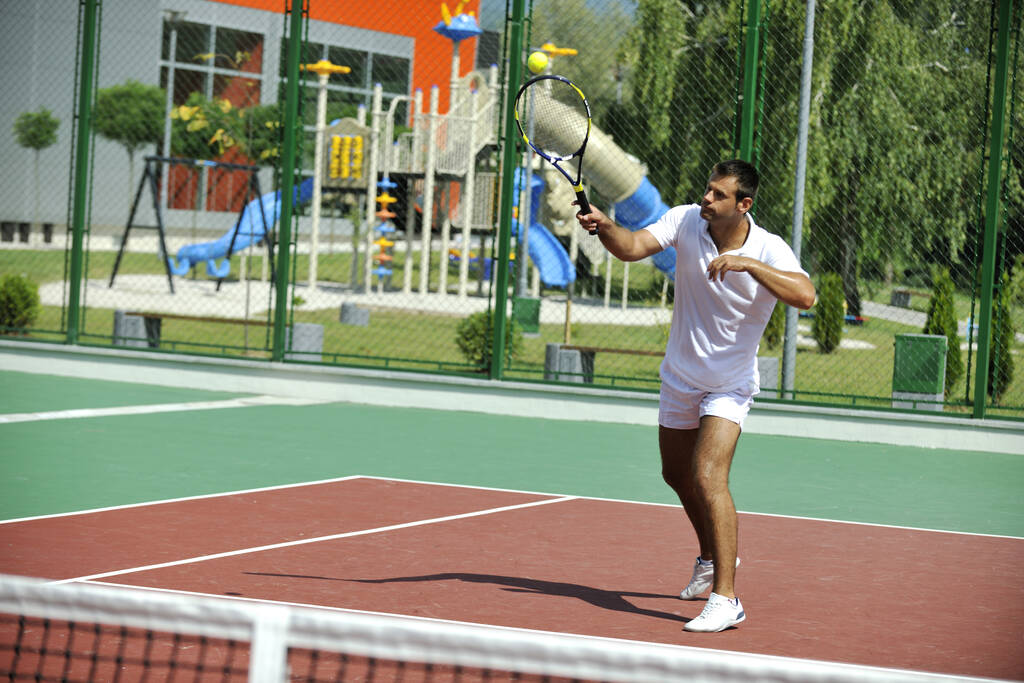 Молодой человек играет в теннис на открытом воздухе на оранжевом теннисном корте рано утром
 - Фото, изображение