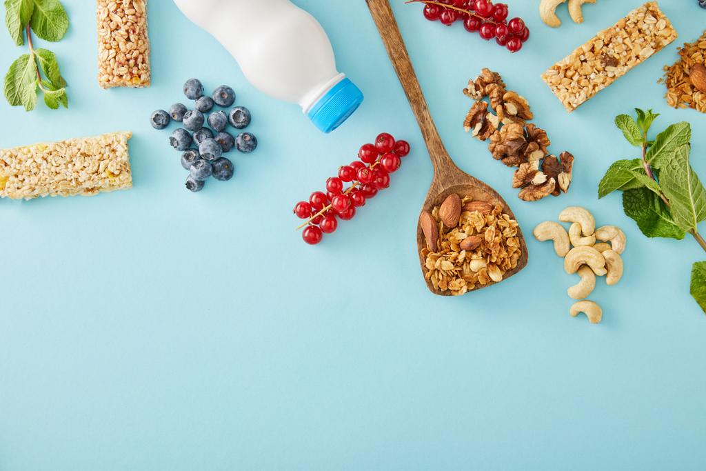 Top view of bottle of yogurt, berries, mint, nuts and cereal bars on blue background - Photo, Image