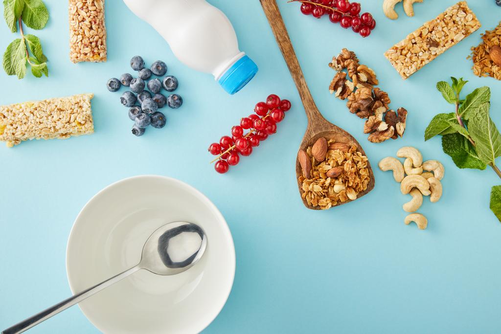 Bovenaanzicht van lepel in lege kom met bessen, munt, noten, fles yoghurt en graanrepen op blauwe achtergrond - Foto, afbeelding