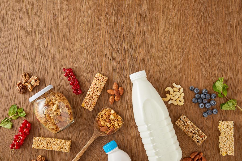 Horní pohled na láhve mléka a jogurtu s bobulemi, mátou, ořechy, cereáliemi a sklenicí granoly na dřevěném pozadí - Fotografie, Obrázek