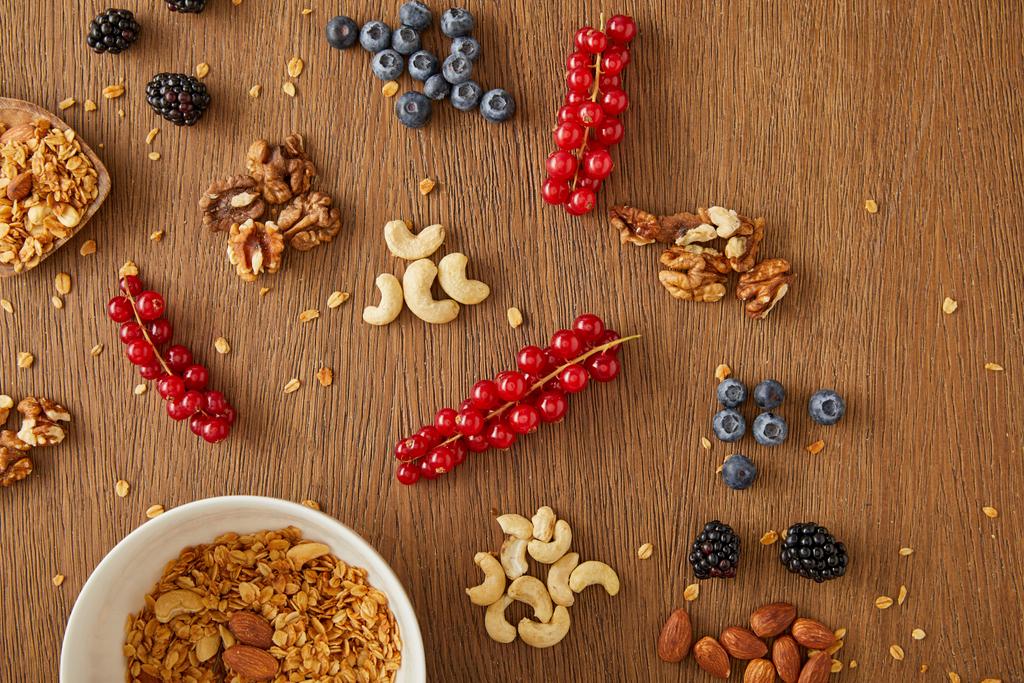 Top view of bowl with granola next to berries, nuts and cereal bars on wooden background - Photo, Image