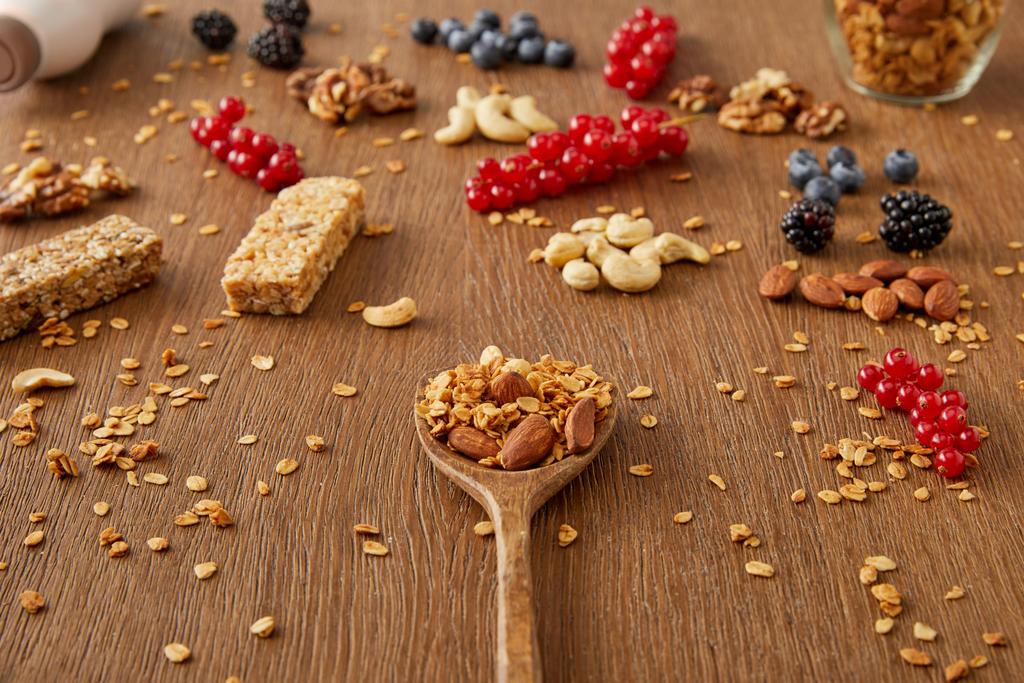Espátula de madera con granola junto a bayas, frutos secos, copos de avena y barras de cereales sobre fondo de madera
  - Foto, imagen