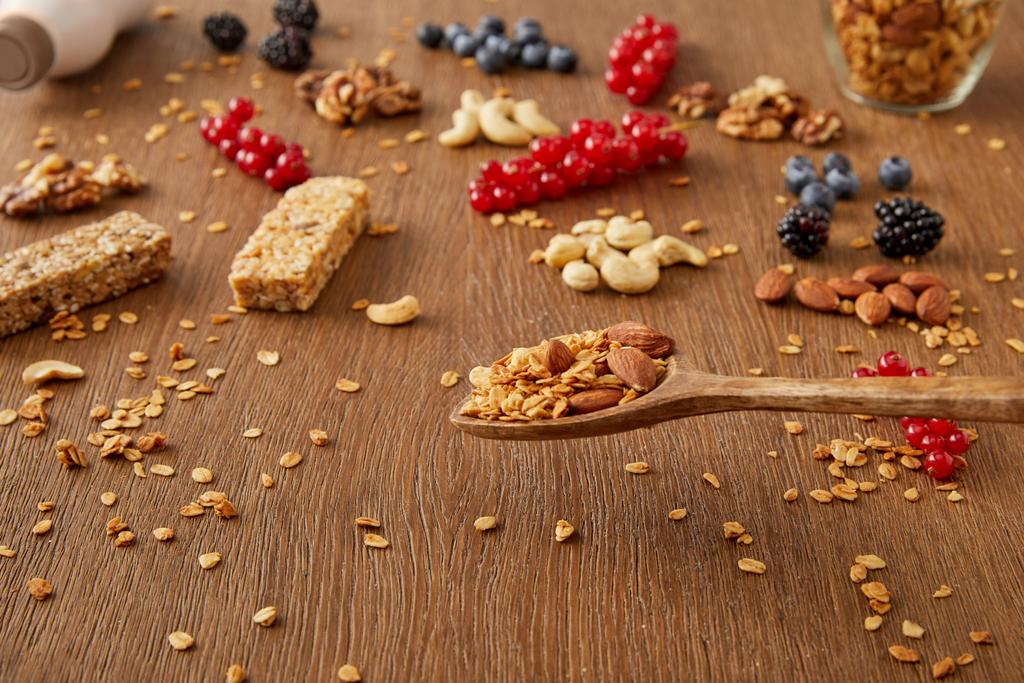 Dřevěná špachtle nad stolem s granolou vedle bobulí, ořechů a obilných tyčinek na dřevěném pozadí - Fotografie, Obrázek