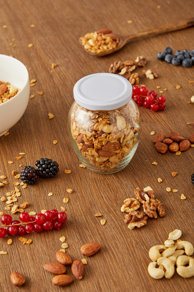 Jar of granola, bowl and spatula next to almonds, walnuts, cashews, berries on wooden background - Photo, Image