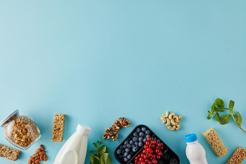 Top view of container with berries, bottles of yogurt and milk, jar of granola, nuts, cereal bars and mint on blue background - Photo, Image