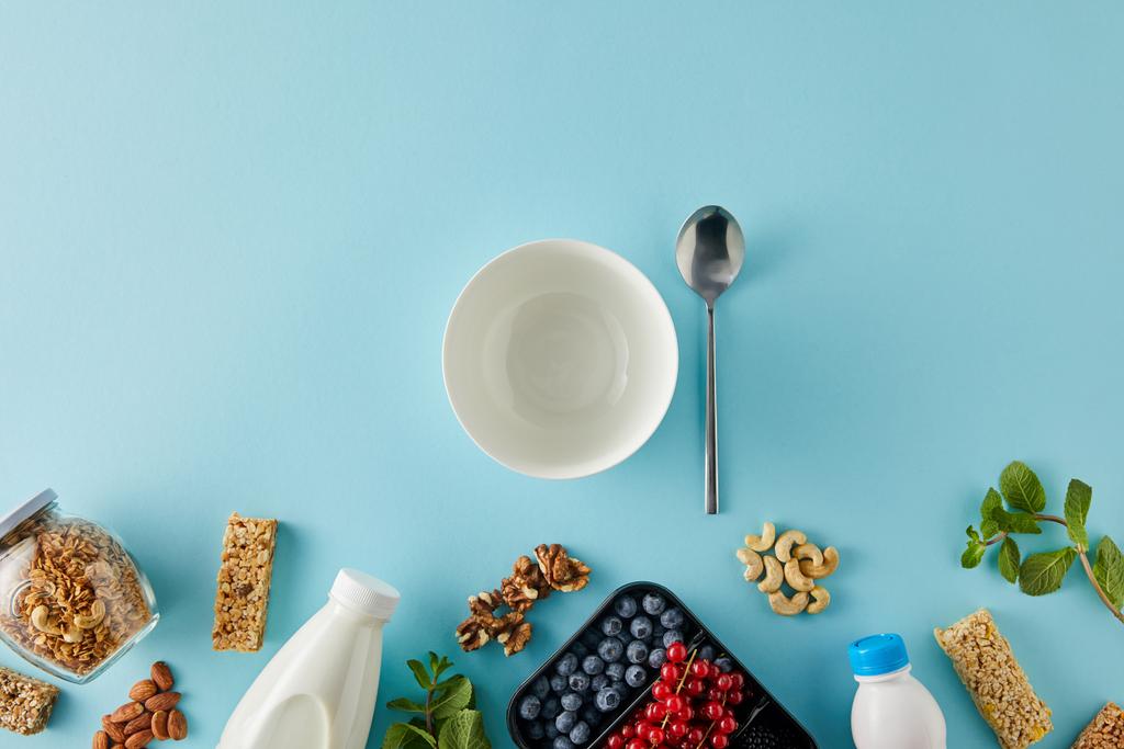 Top view of container with berries, bottles of yogurt and milk, jar of granola, nuts, cereal bars, bowl, spoon on blue background - Photo, Image