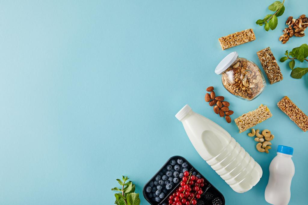 Top view of container with berries, bottles of yogurt and milk, jar of granola, nuts, cereal bars on blue background - Photo, Image