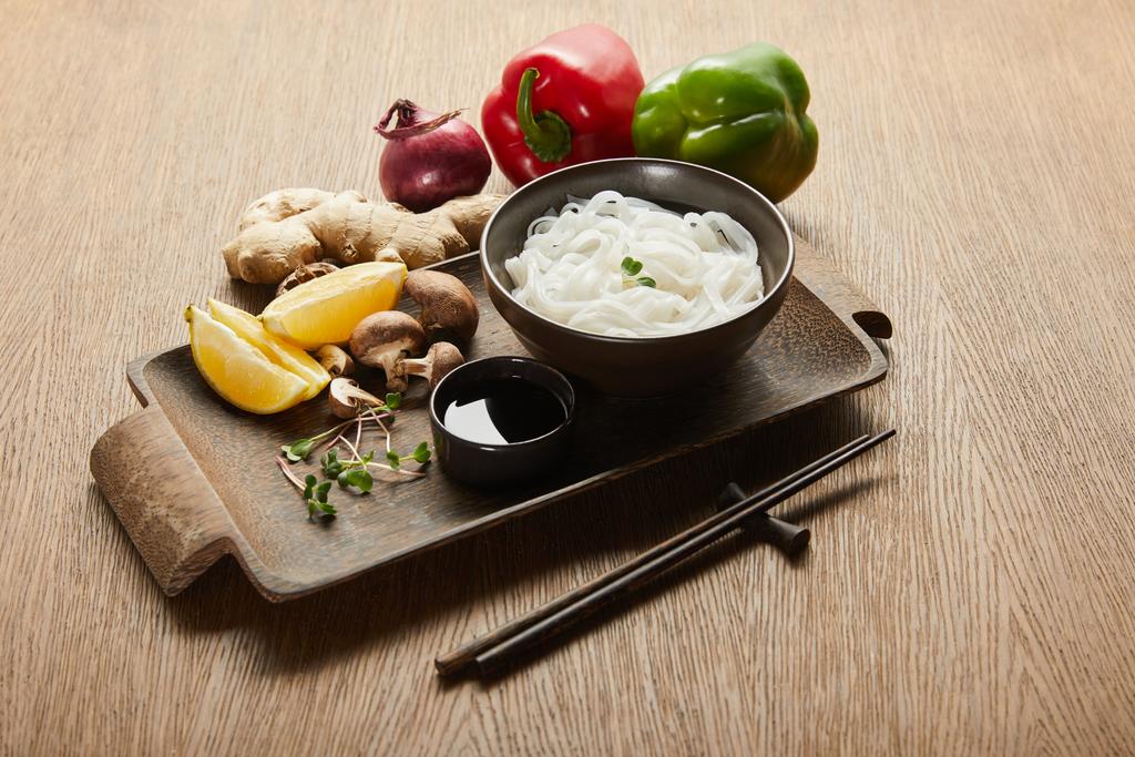 rizs tészta tálban evőpálcikák, szójaszósz, gyömbér gyökér, citrom és zöldség mellett fa tálcán - Fotó, kép