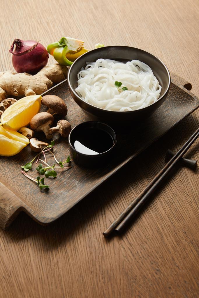 рисовая лапша в миске возле палочек, соевый соус, корень имбиря, лимон и овощи на деревянном подносе
 - Фото, изображение