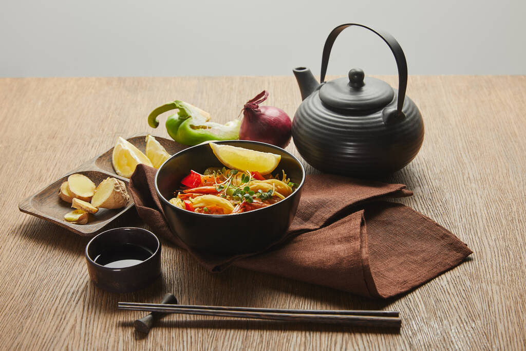 Лапша с креветками и овощами в миске возле палочек, соевый соус, корень лимона и имбиря, чайник на салфетке на деревянном столе, изолированном на сером
 - Фото, изображение
