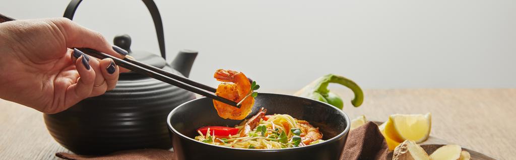 καλλιεργημένη άποψη της γυναίκας κρατώντας γαρίδες με chopsticks κοντά noodles σε μπολ απομονώνονται σε γκρι, πανοραμική λήψη - Φωτογραφία, εικόνα