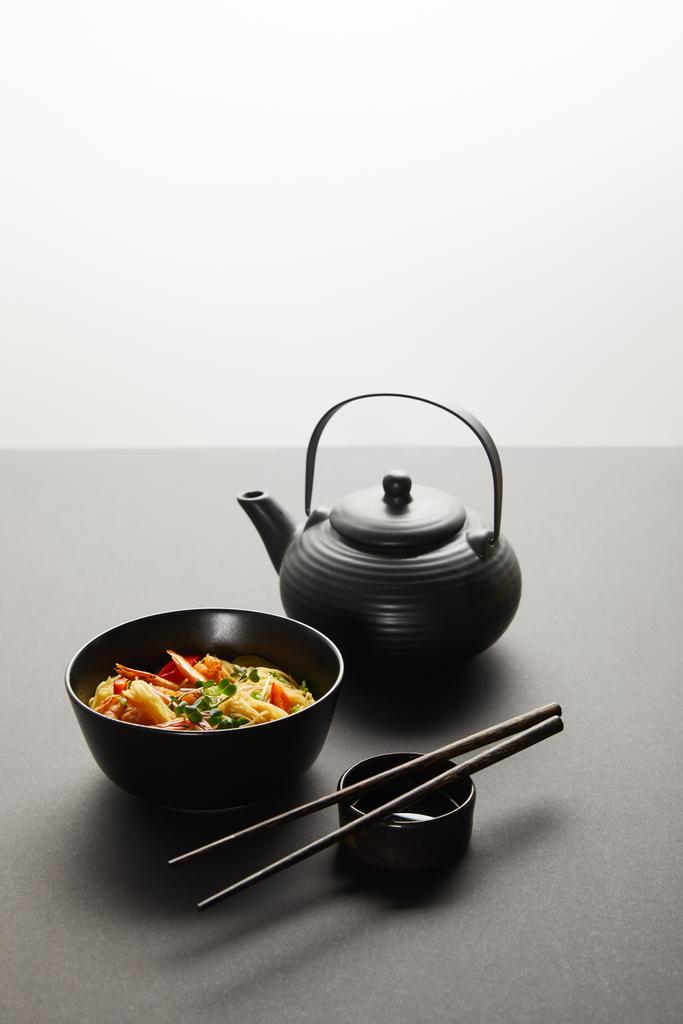 noodles με γαρίδες και λαχανικά σε μπολ κοντά σε λεμόνι, τσαγιέρα, ξύλινα chopsticks και σάλτσα σόγιας σε μαύρο φόντο απομονώνονται σε γκρι - Φωτογραφία, εικόνα