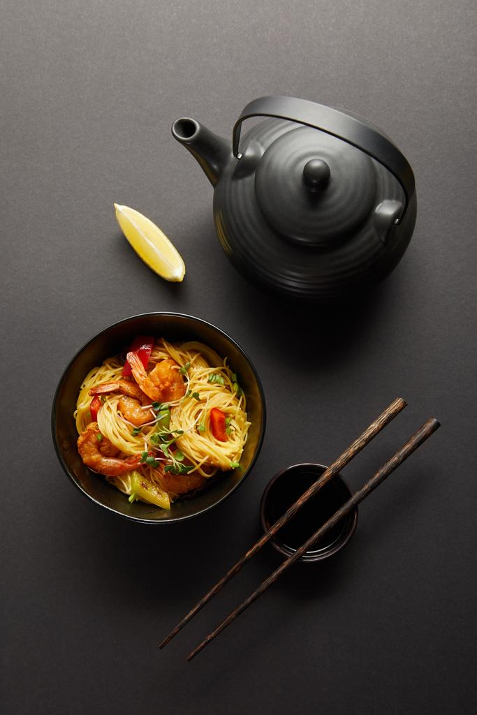 верхний вид лапши с креветками и овощами в миске рядом с лимоном, чайник, деревянные палочки и соевый соус на черном фоне
 - Фото, изображение