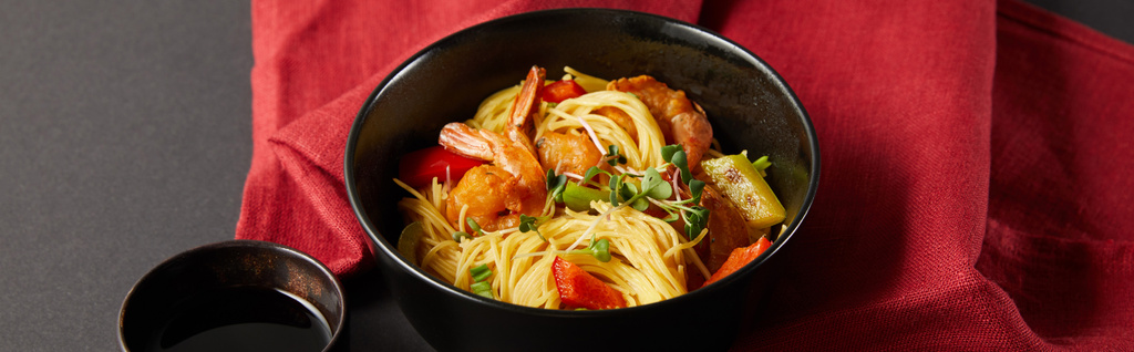 noodles with shrimps and vegetables in bowl near soy sauce on red napkin on black background, panoramic shot - Photo, Image