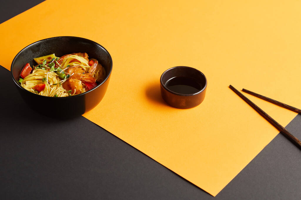 fideos con camarones y verduras en el tazón cerca de los palillos, salsa de soja en fondo negro y amarillo. - Foto, Imagen