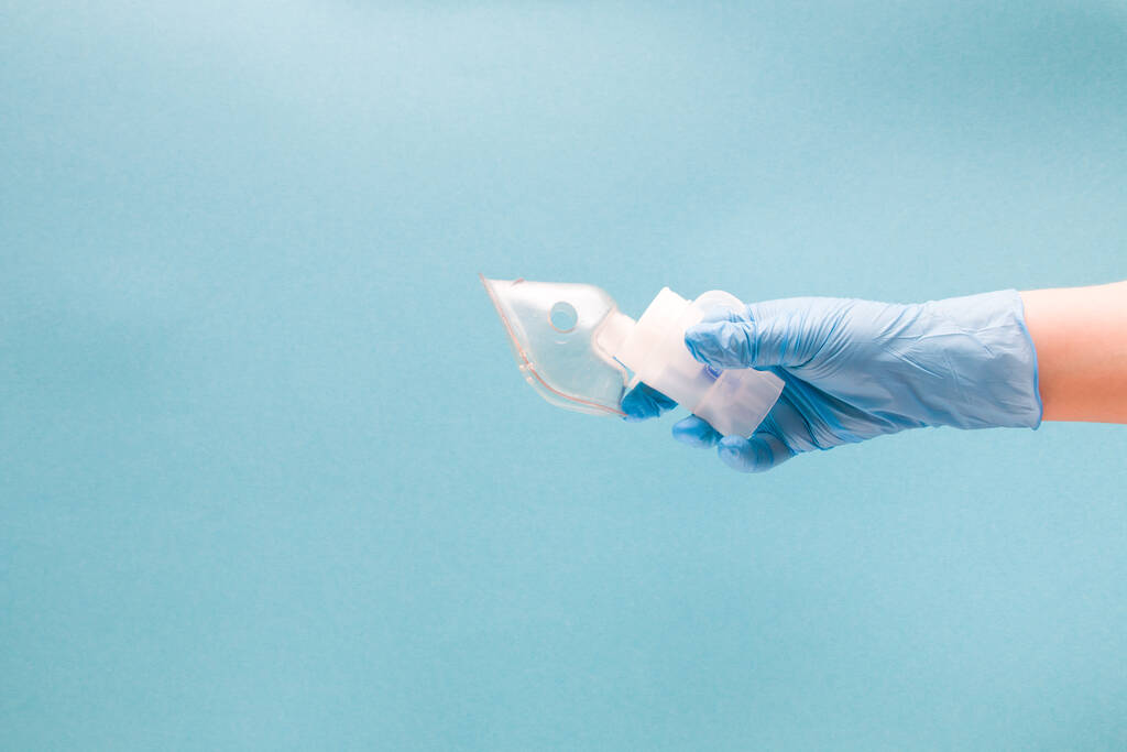 uma mão feminina em uma luva médica descartável azul segura um reservatório de medicação nebulizador com uma máscara de bebê, dispositivo de inalação, conceito de doença do trato respiratório, pneumologia e tratamento de infecções respiratórias, fundo azul, espaço de cópia
 - Foto, Imagem