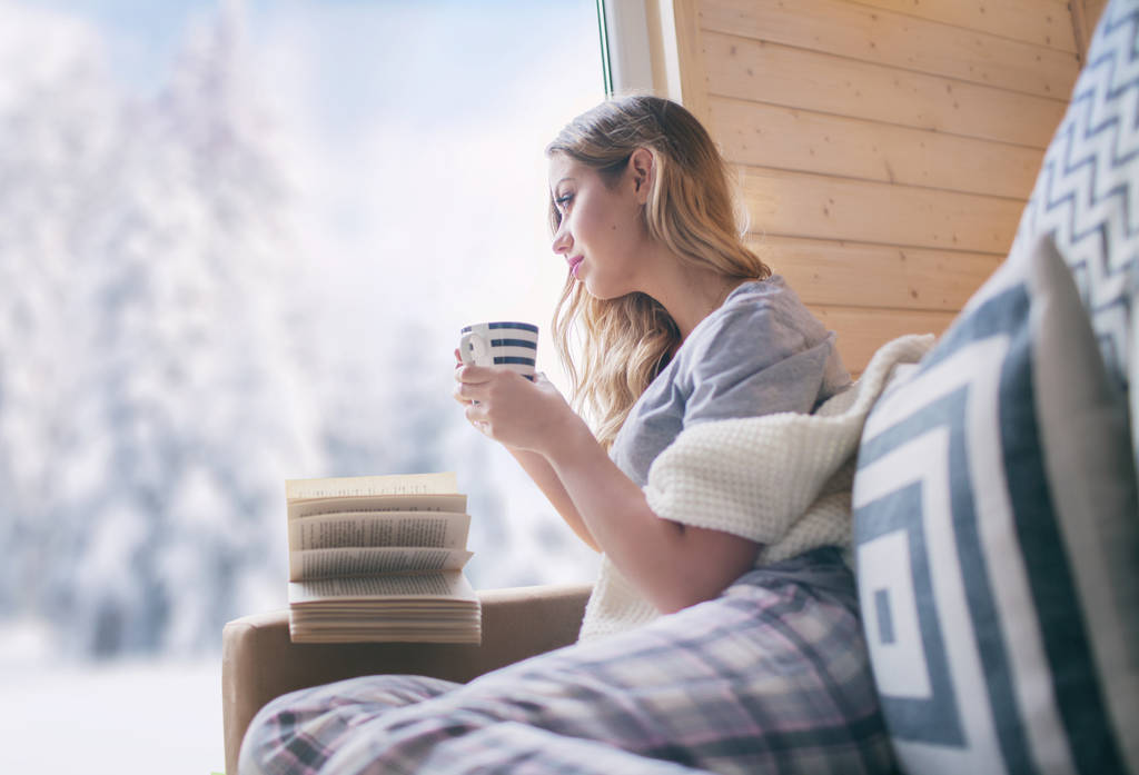 Молодая красивая блондинка с чашкой кофе сидит дома в гостиной у окна. Зимний вид на снег. Концепция Рождества
 - Фото, изображение