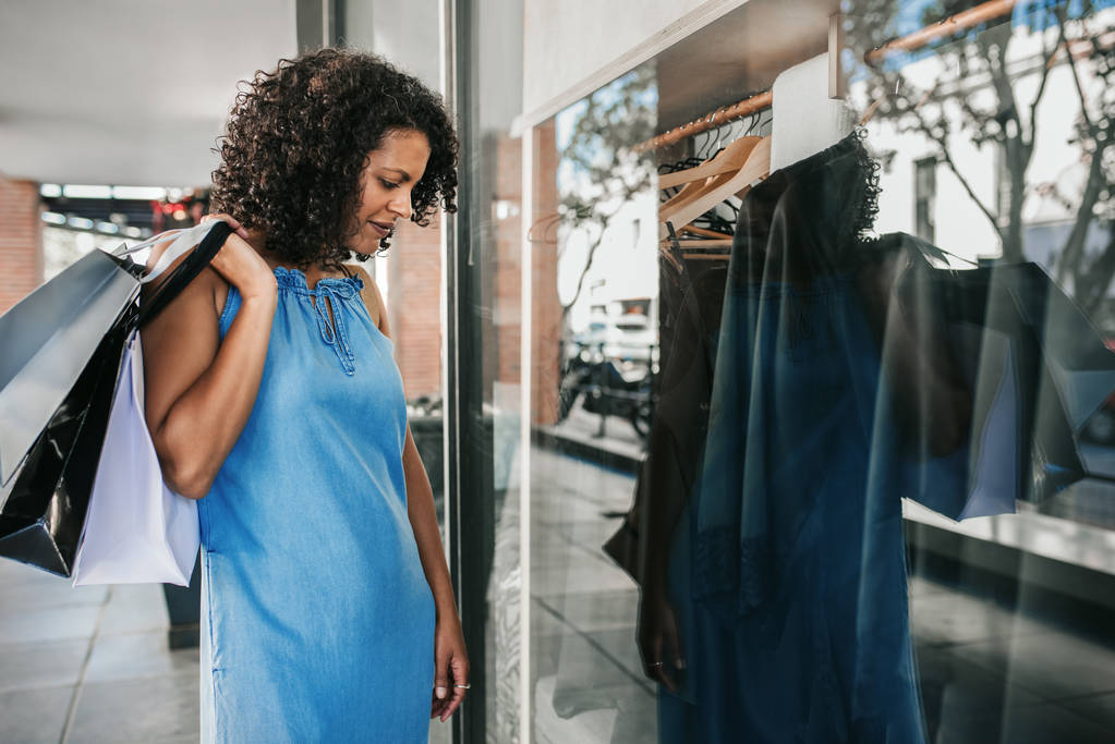Hymyilevä nainen seisoo jalkakäytävällä ja katselee vaatekaupan näyteikkunaa ostoksilla ollessaan
 - Valokuva, kuva