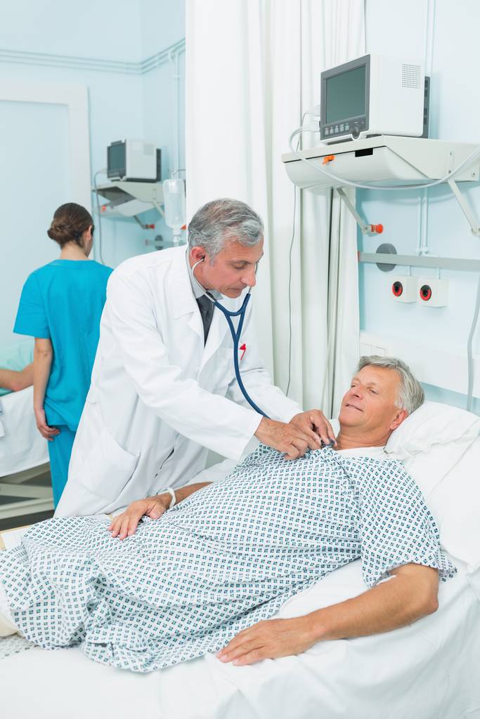 Orvos auscultating egy férfi beteg egy ágyban kórteremben egy kórházban - Fotó, kép