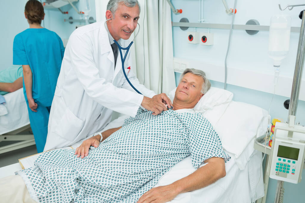 病院のベッド病棟で男性患者の胸を会計検査をする男性医師の笑顔 - 写真・画像