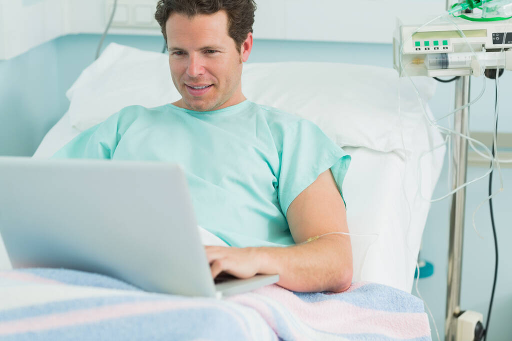 Happy male patient typing on a laptop while lying on a bed in a hospital - Photo, Image