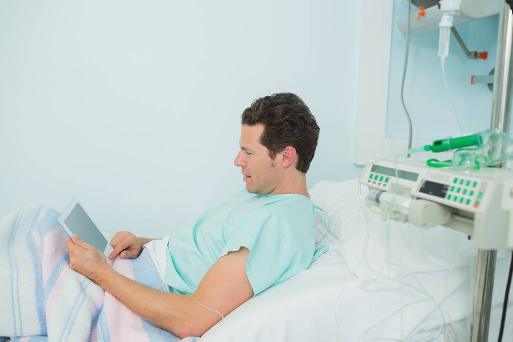 Paciente masculino que toca una tableta táctil mientras se acostaba en una cama de un hospital. - Foto, imagen