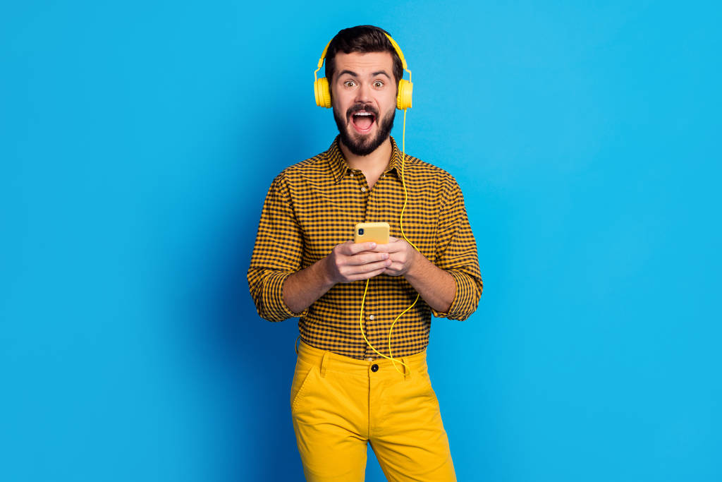 A kedvenc dalom. Megdöbbent izgatott férfi használ smartphone keresés sztereó hangerő dallam sikoly hallgatni zene kopás headset kockás nadrág stílus stílusos trendi elszigetelt kék háttér - Fotó, kép