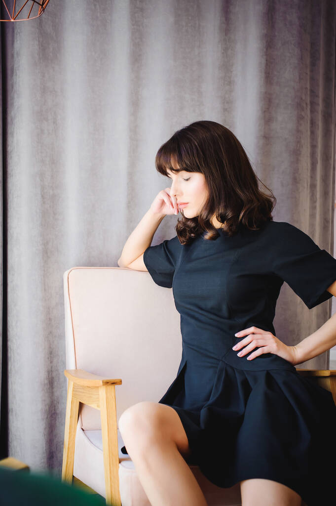 Νεαρή ελκυστική γυναίκα με μακριά πόδια σε μαύρο κομψό φόρεμα, κάθεται στην καρέκλα κοντά στο παράθυρο στο εσωτερικό του δωματίου. - Φωτογραφία, εικόνα