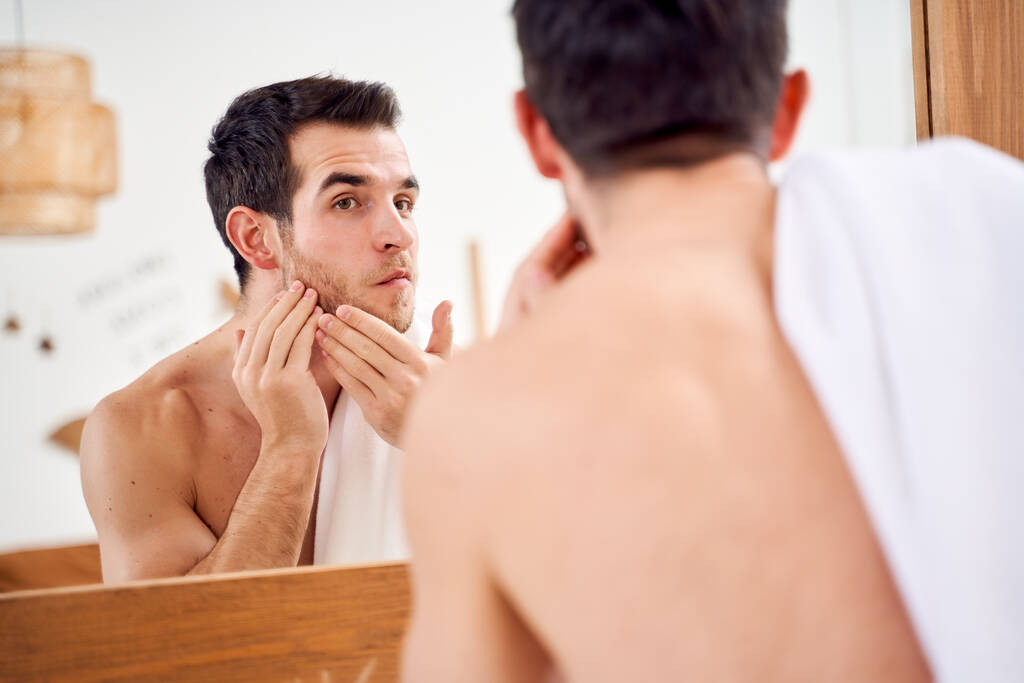 Brunet male lathers his face and stands with towel on his shoulders in front of bathroom mirror - Photo, Image