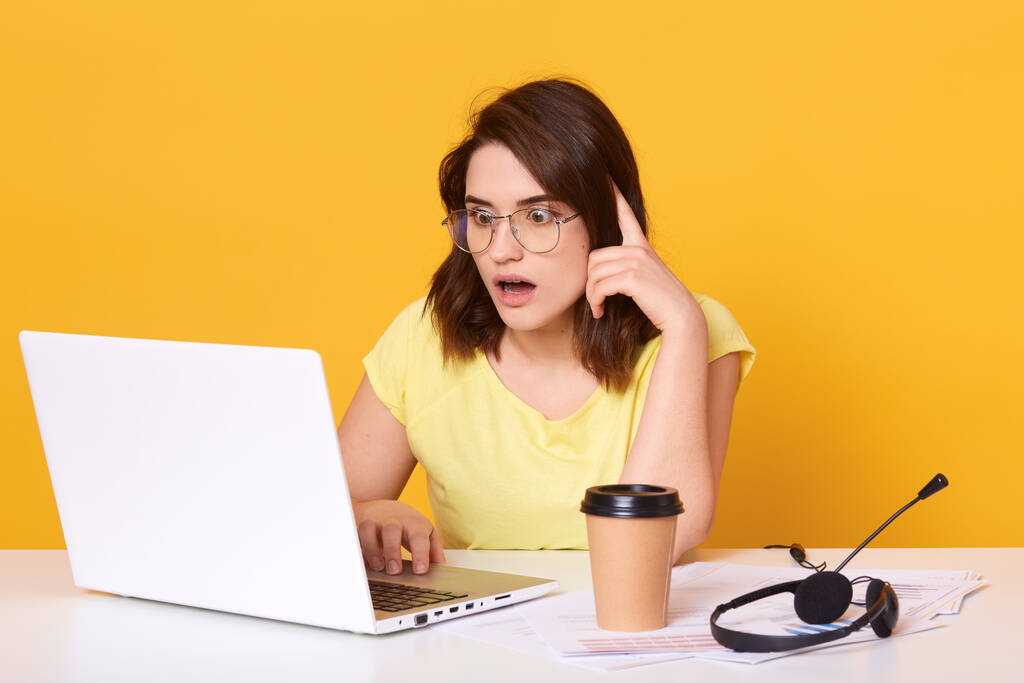 Horizontal shot of business woman works online on her laptop while sitting at desk, looking at screen with widely opened mouth, has astonished and shocked facial expression, isolated over yellow wall - Photo, Image