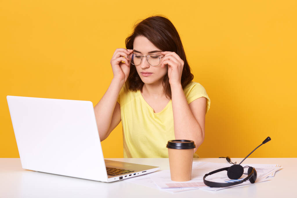 Studyjne ujęcie siedzącej obok białej bluzki i zabierającej kawę, dotykającej jej okularów, wygląda na zmęczoną, pracującą online, uroczą brunetkę pozującą na żółtym tle. Koktajl technologiczny. - Zdjęcie, obraz