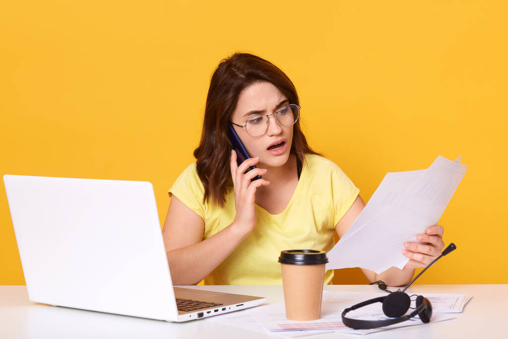 Portret biznesmenki pracującej online przez laptopa, odizolowanej na żółtym tle studia, rozmawiającej z kimś przez smartfon, trzymającej papier w rękach, wygląda zdumiewająco. i skoncentrowany. - Zdjęcie, obraz