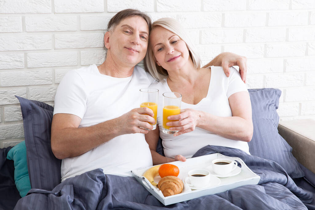 Ευτυχισμένο μεσήλικο ζευγάρι τρώει πρωινό στο κρεβάτι στο ξενοδοχείο - Φωτογραφία, εικόνα